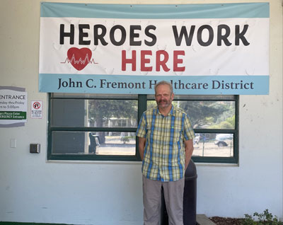 Picture of Robin Fry, John C. Fremont Provider- standing in front of a banner hanging on the building. It says: HEROES WORK HERE (HEART)