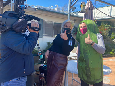 Picture of a man holding a large video camera on his shoulder and two females are wearing mask and looking at the camera, giving a thumbs up.
